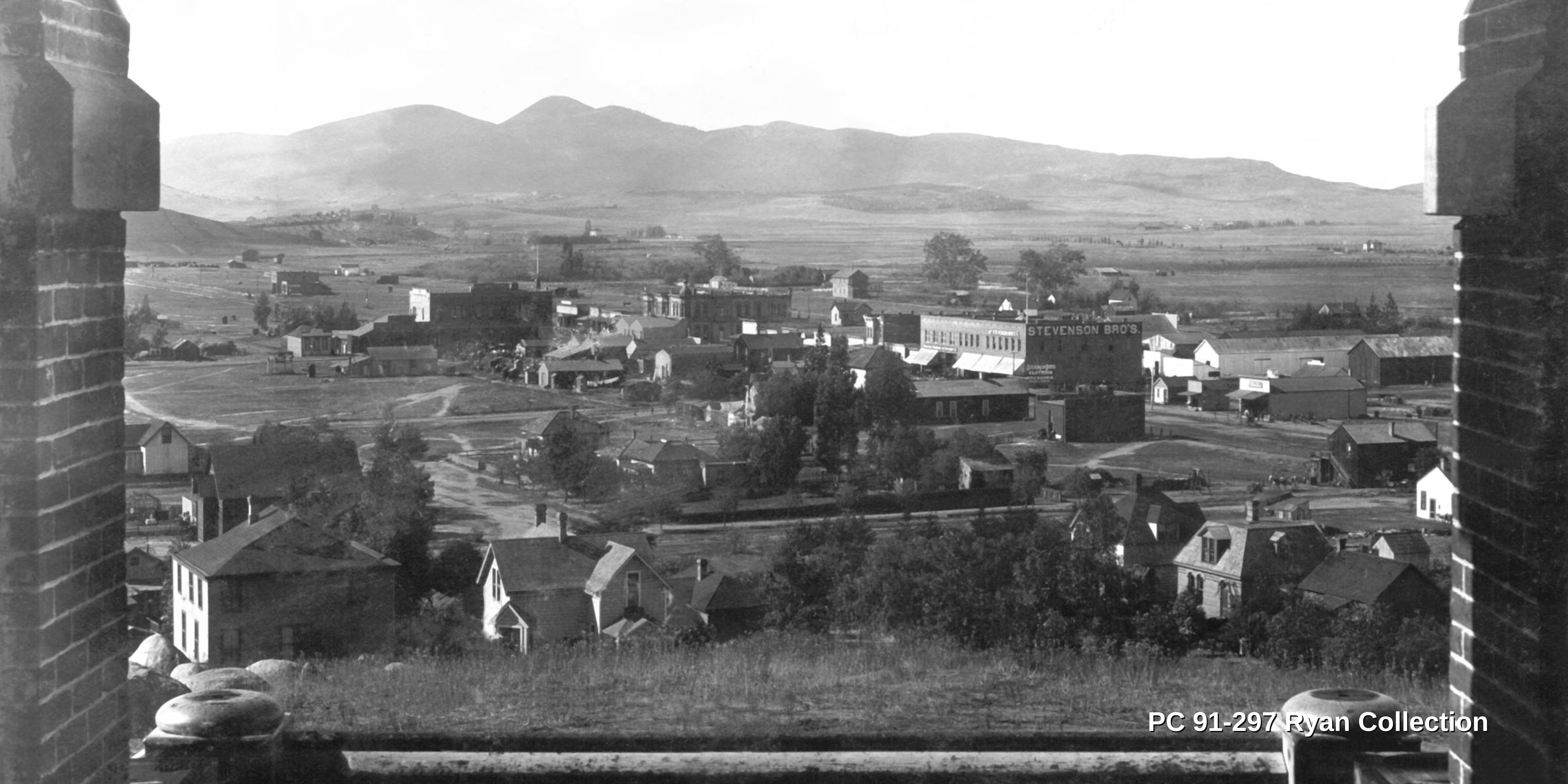 Image of historic Escondido from the Frances Beven Ryan Collection. This black and white photo shows homes and businesses from the balcony of the Escondido High School. There is a view of the mountains in the distance and there are noticeably fewer buildings compared to today's Escondido.